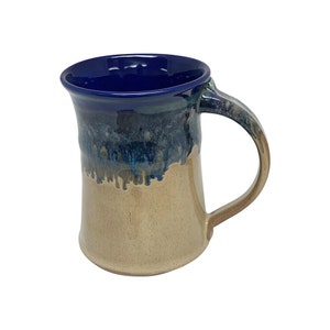 May include: Handmade ceramic mug with a dark blue interior and handle. The exterior transitions from a tan base to a blue and teal glaze, creating a unique, dripped effect. The mug has a large handle for easy gripping.
