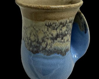 May include: A handmade ceramic mug with a unique color gradient. The top rim is a light brown, transitioning to a speckled tan and grey mid-section, and finishing with a solid blue base and handle. The mug has a curved handle and a wide mouth.