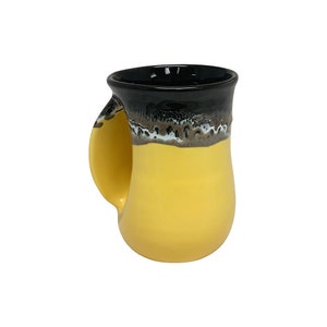 May include: A yellow ceramic mug with a black rim and a unique handle that wraps around the side. The mug has a brown and white glaze pattern around the rim.