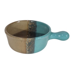 May include: A small, round ceramic bowl with a handle. The bowl is glazed in a light brown color on one side and a turquoise color on the other side. The handle is also turquoise.