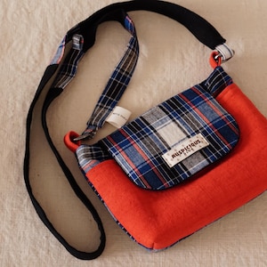 May include: A red and plaid crossbody bag with a black strap. The bag features a flap with a blue, white, and red plaid pattern. The bag has a tag that reads "AUSPICIOUS KIDS".