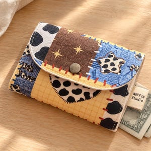 May include: A bifold wallet featuring a patchwork design of cow print, yellow grid patterns, and brown fabric with yellow stars. The flap has a metal snap closure and a star-shaped patch. A small tag reads MUFFIN Studio, with paper currency visible.
