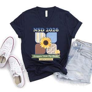 May include: Navy blue V-neck t-shirt with a sunflower and chick graphic. The shirt features the text "NSD 2026" and "Scrappin' With My Chicks". The shirt is paired with denim shorts and white sneakers.