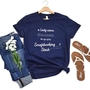 May include: A navy blue t-shirt with white text that reads "A Lady never discusses the size of her Scrapbooking Stash". The text is accompanied by a small white heart and scissors graphic.