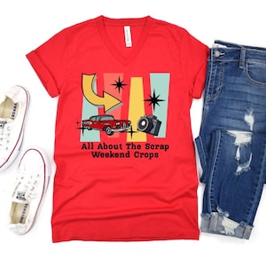 May include: Red V-neck t-shirt with a retro design featuring a red car, camera, and arrow, with the text "All About The Scrap Weekend Crops." The shirt is paired with blue jeans and white sneakers.