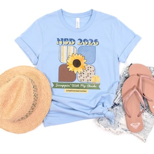 May include: A light blue t-shirt with a sunflower and the text "NSD 2026" and "Scrappin' With My Chicks". A straw hat and pink flip-flops are next to the shirt.
