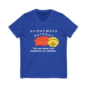 May include: A royal blue V-neck t-shirt with the words "SCRAPBOOK RETREAT" in a stylized font. Below the text is an orange couch graphic. The shirt also features the text "THE ONE WHERE THEY SCRAPBOOK ALL WEEKEND". A yellow hexagon reads "GET YOUR CROP ON" and "CAMP MANITOQUA 2024".