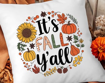 Puede incluir: Cojín cuadrado blanco con un diseño de temática otoñal. Presenta una disposición circular de un girasol, calabazas, hojas y el texto "It's FALL y'all" en varios colores. El cojín está colocado sobre una silla de madera con una manta marrón.