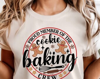 May include: A cream-coloured t-shirt featuring a circular graphic that says "Proud Member of the Cookie Baking Crew." The design includes gingerbread men, snowflakes, stars, a whisk, and a rolling pin. The shirt is being worn.