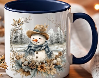 May include: A white ceramic mug with a dark blue interior and handle, featuring a printed illustration of a snowman wearing a hat and scarf, surrounded by a wreath of dried flowers, pinecones, and foliage against a winter forest background.