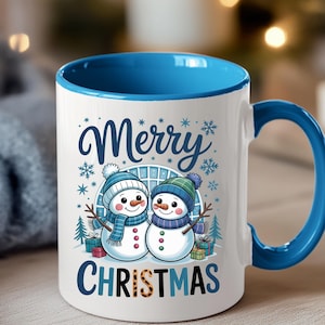Könnte beinhalten: Weißer Keramikbecher mit blauem Innenraum und Henkel. Der Becher hat ein festliches Design mit zwei Schneemännern mit Hüten und Schals, mit dem Aufdruck "Merry Christmas" auf der Vorderseite.