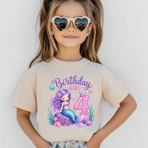 May include: A beige short-sleeved t-shirt featuring a graphic print of a purple-haired mermaid sitting on a rock, surrounded by sea life and the text Birthday GIRL with a large pink number 4. The design includes pearls, starfish, and shells.