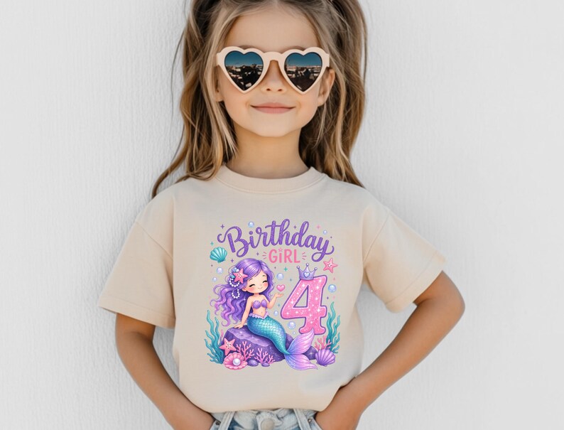 May include: A beige short-sleeved t-shirt featuring a graphic print of a purple-haired mermaid sitting on a rock, surrounded by sea life and the text Birthday GIRL with a large pink number 4. The design includes pearls, starfish, and shells.