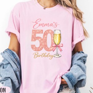 Peut inclure: T-shirt rose clair avec l'inscription "Emma's 50th Birthday". Le chiffre 50 est en rose pailleté, avec un verre de champagne et un nœud rose. Le mot "Birthday" est en or.