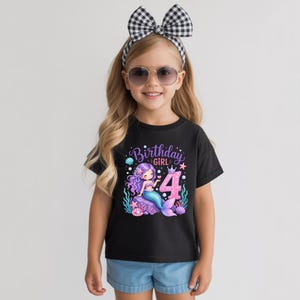 May include: A black short-sleeved t-shirt featuring a graphic of a purple-haired mermaid sitting on a rock, the number 4, and the text Birthday GIRL. The design includes sea elements like shells, starfish, and seaweed in pink and blue tones.