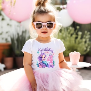 May include: A white short-sleeved t-shirt featuring a graphic of a purple-haired mermaid sitting on a rock next to the number 4 and the text Birthday Girl, paired with a light pink layered tulle tutu skirt and pink-framed sunglasses.