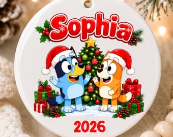 May include: A round white ceramic Christmas ornament featuring the name Sophia in red text at the top. Below, two cartoon dogs wearing Santa hats stand in front of a decorated Christmas tree with presents. The year 2026 is printed in red at the bottom.