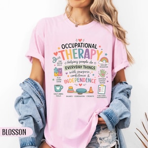 May include: A light pink t-shirt featuring the text OCCUPATIONAL THERAPY Helping people do EVERYDAY THINGS with purpose, confidence & INDEPENDENCE. The design includes colorful icons like scissors, a puzzle piece, a coffee mug, a rainbow, a hand, and a clipboard.