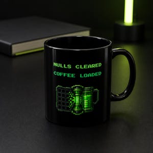 May include: A black ceramic mug featuring the green pixelated text NULLS CLEARED COFFEE LOADED. Below the text is a graphic of a digital loading bar and a battery icon, both rendered in shades of bright green against the dark mug surface.