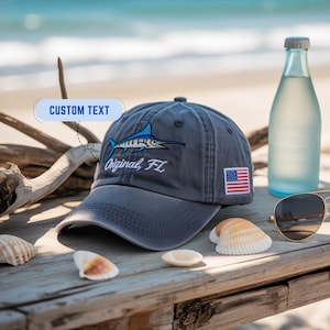 May include: A gray baseball cap with a blue marlin design and the text "Original, FL". The cap features an American flag patch and is displayed with seashells, sunglasses, and a water bottle.