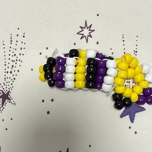 May include: A handmade bracelet crafted from plastic pony beads in a repeating pattern of yellow, white, purple, and black. The beads are strung together on white elastic cord, forming a structured, multi-row band with a textured surface.