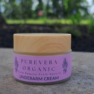 Puede incluir: Un tarro de crema para axilas Purevera Organic con tapa de madera. La etiqueta es lavanda con el texto "Purevera Organic" y "Underarm Cream". El fondo est&aacute; borroso con follaje verde y cielo azul.