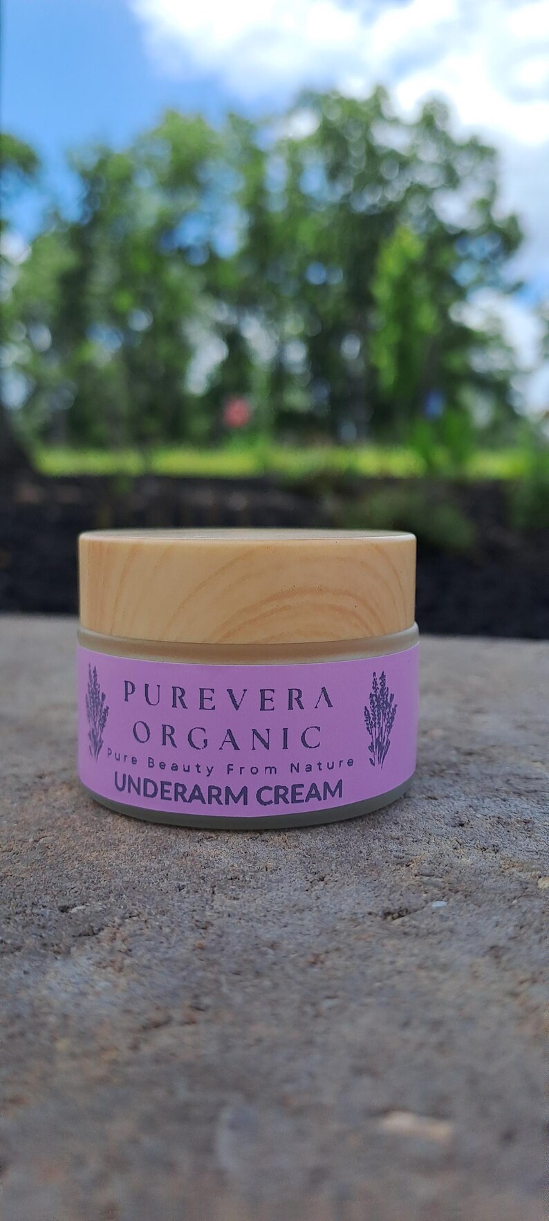 Puede incluir: Un tarro de crema para axilas Purevera Organic con tapa de madera. La etiqueta es lavanda con el texto "Purevera Organic" y "Underarm Cream". El fondo est&aacute; borroso con follaje verde y cielo azul.