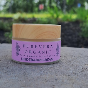 Puede incluir: Un peque&ntilde;o frasco de crema para axilas PUREVERA ORGANIC con tapa de madera. La etiqueta es lavanda con texto e ilustraciones de ramitas de lavanda. El fondo es follaje verde borroso.