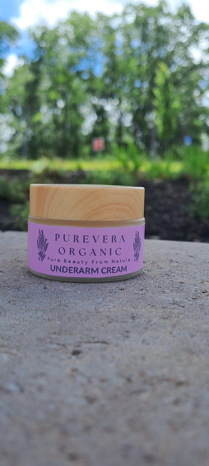 Puede incluir: Un peque&ntilde;o frasco de crema para axilas PUREVERA ORGANIC con tapa de madera. La etiqueta es lavanda con texto e ilustraciones de ramitas de lavanda. El fondo es follaje verde borroso.