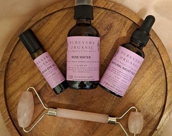 May include: A wooden tray holds skincare products: a rose quartz face roller, a roller bottle, two glass bottles with droppers, and a spray bottle. The bottles have pink labels with black text. The products are from Purevera Organic.