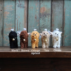 Może przedstawiać: Pięć pluszowych ozdób w kształcie psa anioła w różnych kolorach: czarnym, brązowym, pomarańczowym, brązowym i kremowym. Każda ozdoba ma białą aureolę i skrzydła oraz mały dzwonek wiszący na szyi.