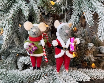 Op de afbeelding: Twee kerstboomversieringen in de vorm van muizen. Eén muis draagt een groene trui, een rode sjaal en houdt een cadeau en een zuurstok vast. De andere is verkleed als kerstman en houdt een roze cadeau vast. Beide hebben witte oren, op een besneeuwde boom.