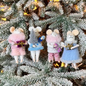 Op de afbeelding: Vier feestelijke muisornamenten hangen aan een met sneeuw bedekte kerstboom. Elke muis draagt een andere outfit, waaronder roze, blauwe en witte jurken, en houdt een cadeau of een miniatuur kerstboom vast. De ornamenten hebben bruine oren en witte gezichten.