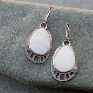 May include: A pair of silver-tone earrings with a teardrop shape. Each earring features a white, oval-shaped stone set within a decorative frame. One earring has a small, purple accent stone. The earrings are displayed on a gray stone surface.