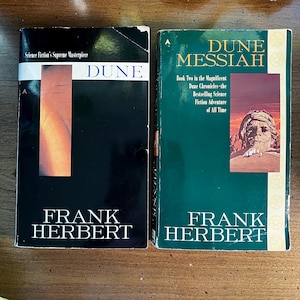 May include: Two books by Frank Herbert. The first, "Dune," has a black cover with a vertical orange and tan design. The second, "Dune Messiah," has a green cover with an illustration of a person. Both books are science fiction.