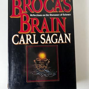 May include: A hardback book titled "Broca's Brain" by Carl Sagan. The title is in large red letters with gold trim, set against a black background. The cover features an illustration of a face with a glowing aura.