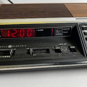Könnte beinhalten: Ein Vintage General Electric Digital-Radiowecker mit Holzmaserung und schwarzem Bedienfeld. Die rote Digitalanzeige zeigt 12:00 Uhr an. Funktionen umfassen Helligkeit, Lautstärke und Radiosteuerung.