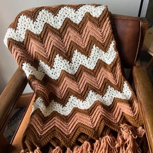 Puede incluir: Una manta de ganchillo hecha a mano con un patr&oacute;n de chevr&oacute;n en tonos marr&oacute;n, crema y melocot&oacute;n. La manta est&aacute; sobre una silla de cuero marr&oacute;n y tiene una franja de hilo color melocot&oacute;n en el borde inferior.