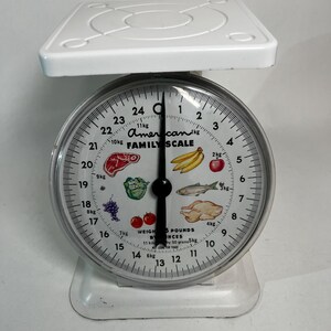 May include: A vintage white American Family Scale with a circular dial. The dial features illustrations of food items and weight measurements in pounds and kilograms. The scale has a platform on top and a base.