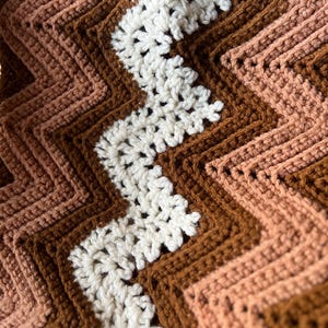 Puede incluir: Primer plano de una manta de ganchillo hecha a mano con un patr&oacute;n de chevr&oacute;n. La manta presenta franjas alternas de blanco, marr&oacute;n claro, marr&oacute;n medio y marr&oacute;n oscuro. La textura del hilo es visible, creando una apariencia acogedora y c&aacute;lida.