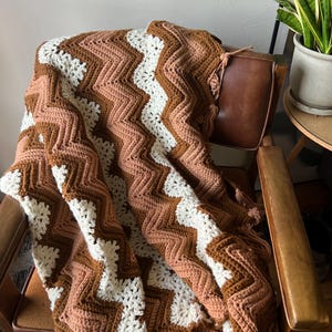 Puede incluir: Una manta de ganchillo hecha a mano, sobre una silla de cuero marr&oacute;n. La manta presenta un patr&oacute;n de chevr&oacute;n en tonos marr&oacute;n, crema y melocot&oacute;n. La manta tiene un borde con flecos. La silla est&aacute; en una habitaci&oacute;n con una planta.