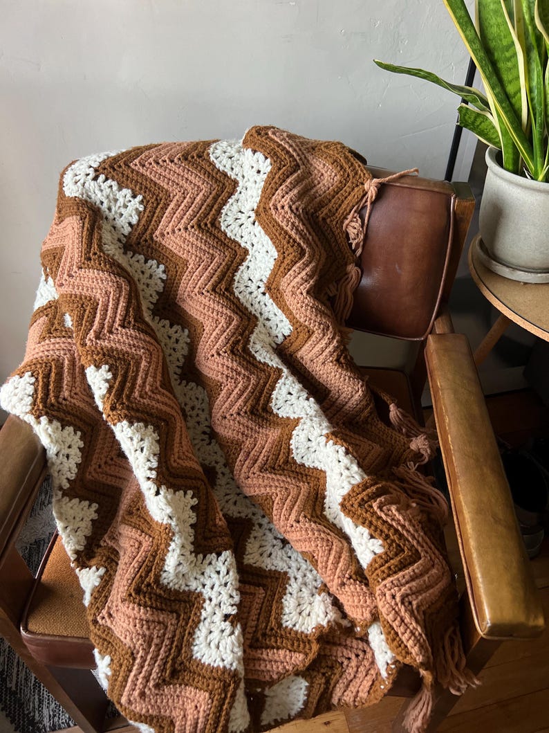 Puede incluir: Una manta de ganchillo hecha a mano, sobre una silla de cuero marr&oacute;n. La manta presenta un patr&oacute;n de chevr&oacute;n en tonos marr&oacute;n, crema y melocot&oacute;n. La manta tiene un borde con flecos. La silla est&aacute; en una habitaci&oacute;n con una planta.