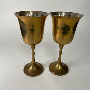 Puede incluir: Dos copas vintage de latón con borde acampanado y tallo delgado. Las copas tienen un acabado dorado pulido y un interior plateado. Las copas se exhiben sobre un fondo blanco.