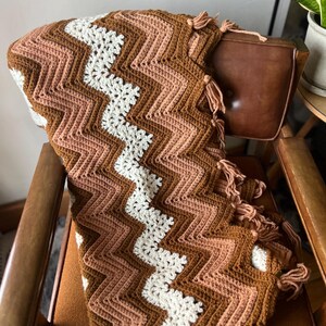 Puede incluir: Una manta de ganchillo hecha a mano con un patr&oacute;n de chevr&oacute;n en tonos marr&oacute;n, melocot&oacute;n y blanco. La manta est&aacute; colocada sobre una silla y tiene borlas en un borde. Un textil para el hogar acogedor y decorativo.