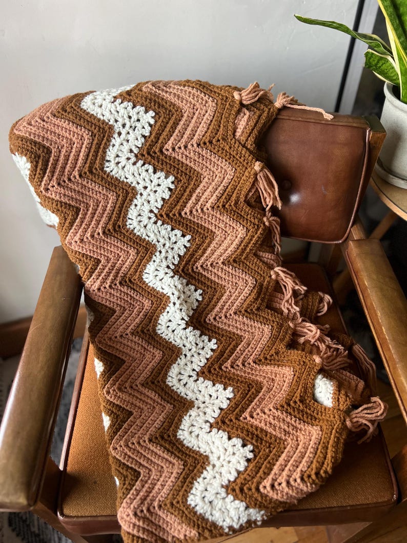 Puede incluir: Una manta de ganchillo hecha a mano con un patr&oacute;n de chevr&oacute;n en tonos marr&oacute;n, melocot&oacute;n y blanco. La manta est&aacute; colocada sobre una silla y tiene borlas en un borde. Un textil para el hogar acogedor y decorativo.