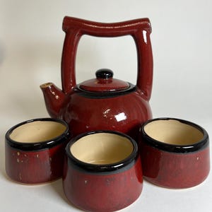 Könnte beinhalten: Rotes Keramik-Teeservice mit schwarzem Deckel und Griff, sowie drei passenden Tassen. Die Teekanne und Tassen haben eine glänzende Oberfläche und einen schwarzen Rand. Die Tassen haben einen beigefarbenen Innenraum.