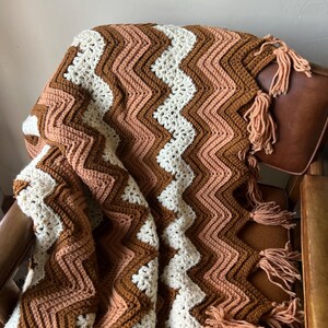 Puede incluir: Una manta de ganchillo hecha a mano con un patr&oacute;n de chevr&oacute;n en tonos marr&oacute;n, crema y melocot&oacute;n. La manta tiene borlas y est&aacute; colocada sobre una silla. Mide aproximadamente 150 cm x 200 cm. Ideal para a&ntilde;adir calidez y estilo a cualquier habitaci&oacute;n.