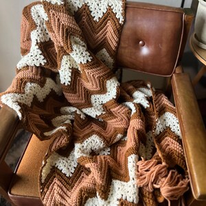 Puede incluir: Una manta de ganchillo hecha a mano con un patr&oacute;n de chevr&oacute;n en tonos marr&oacute;n, crema y melocot&oacute;n. La manta est&aacute; sobre una silla vintage de cuero marr&oacute;n. Los flecos adornan el borde de la manta, a&ntilde;adiendo un toque decorativo.