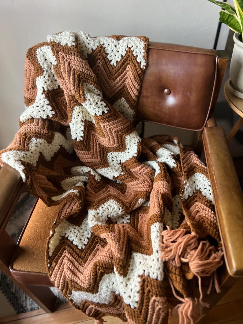 Puede incluir: Una manta de ganchillo hecha a mano con un patr&oacute;n de chevr&oacute;n en tonos marr&oacute;n, crema y melocot&oacute;n. La manta est&aacute; sobre una silla vintage de cuero marr&oacute;n. Los flecos adornan el borde de la manta, a&ntilde;adiendo un toque decorativo.