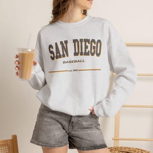 Puede incluir: Una sudadera gris claro de cuello redondo con el texto SAN DIEGO BASEBALL en una fuente oscura desgastada con bordes amarillos. Debajo del texto hay una línea horizontal y la frase est. 1969. La sudadera tiene mangas largas y cuello acanalado.